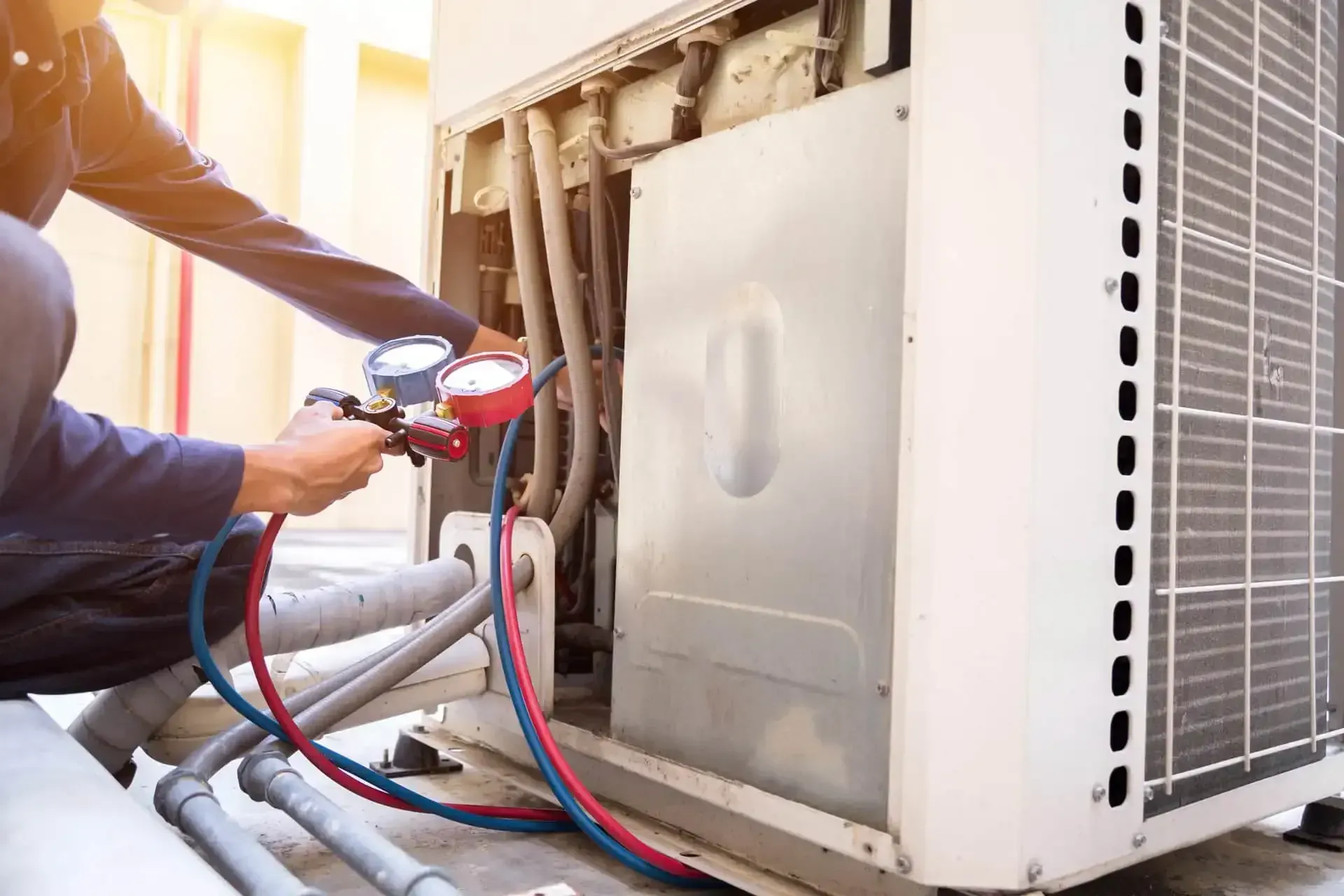 Technician checking refrigerant pressure with manifold gauges on an outdoor AC condenser.