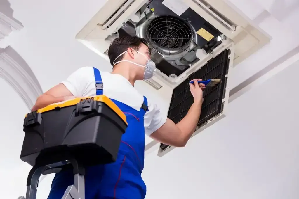 Technician on a ladder cleaning the filter of a ceiling-mounted air conditioning unit.