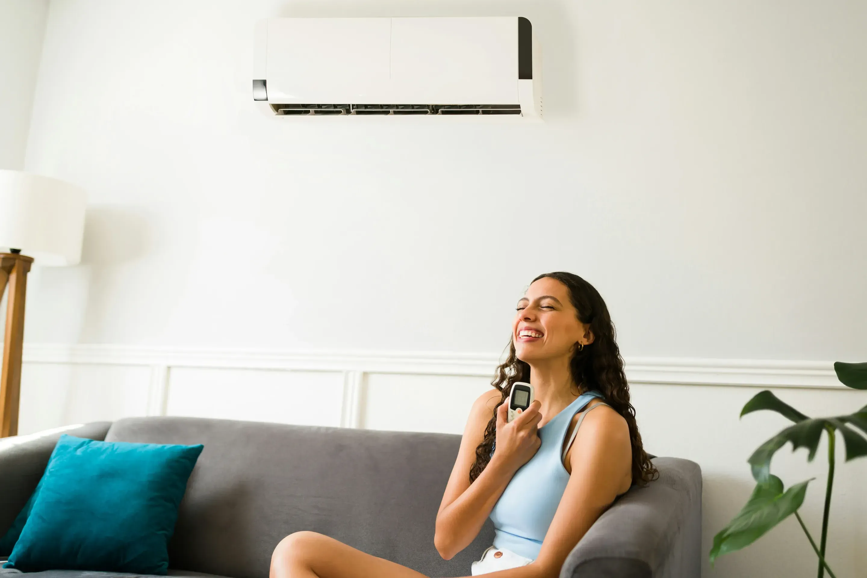 Smiling woman on a sofa holding an AC remote beneath a wall-mounted mini-split air conditioner.